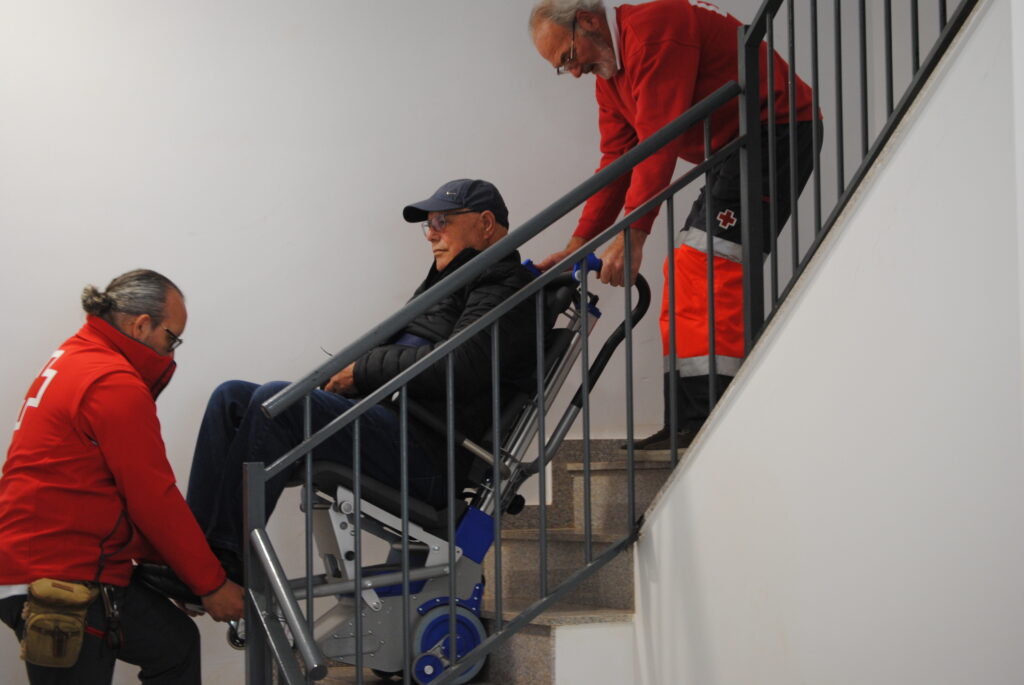 Deux bénévoles de la Croix-Rouge portent une personne âgée dans les escaliers.