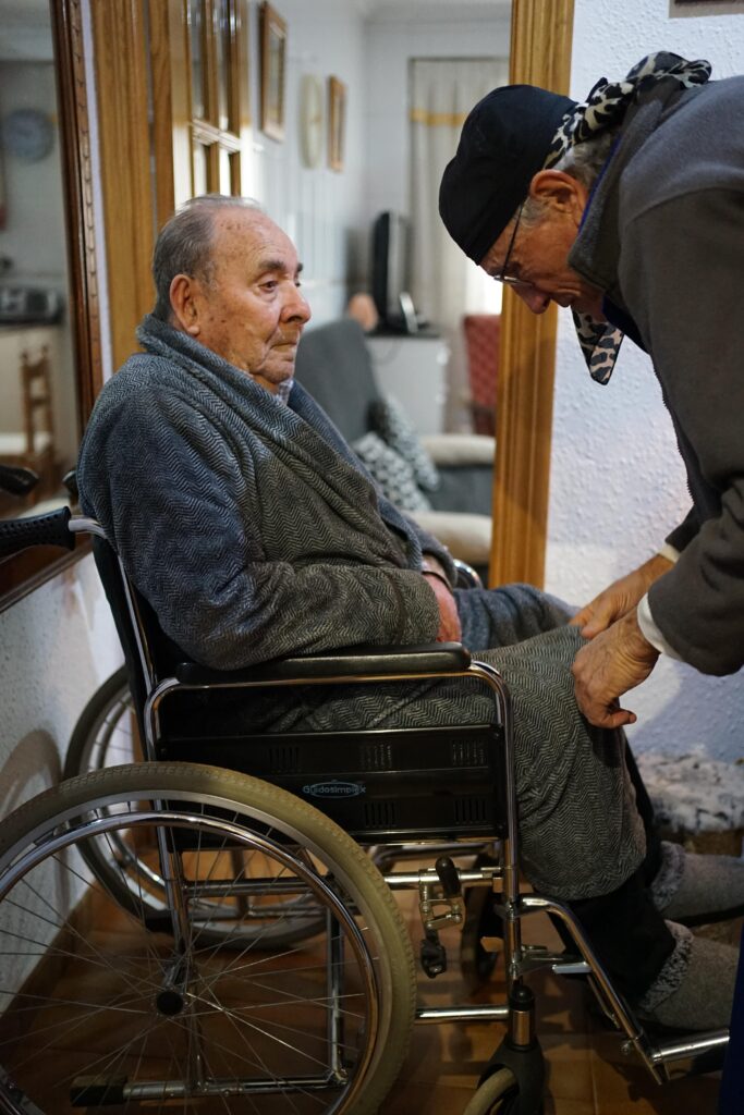 Un bénévole aide une personne âgée à s'installer sur son fauteuil roulant.